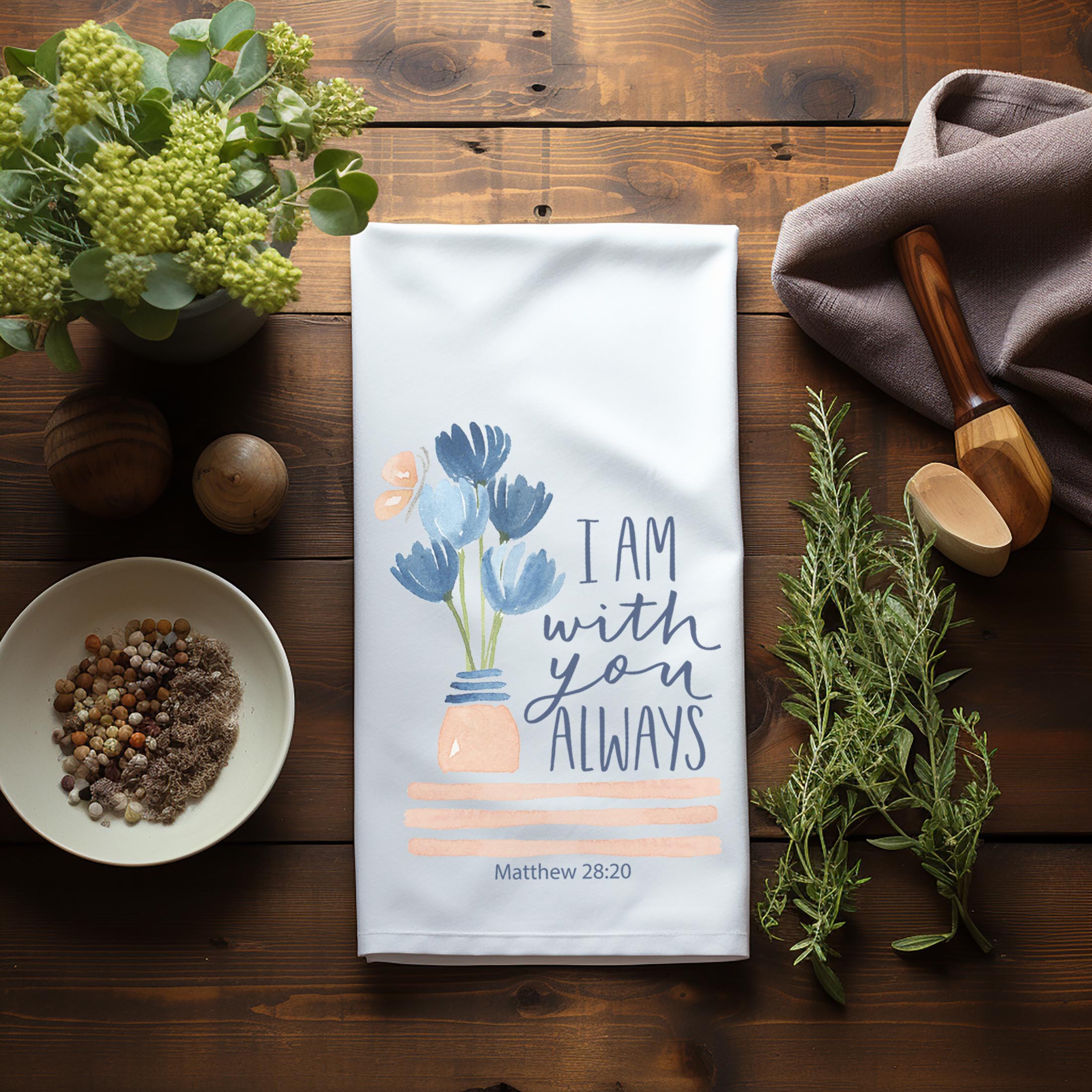 Christian tea towel with floral design and text "I Am With you Always" on a wooden surface with plants and kitchen items.