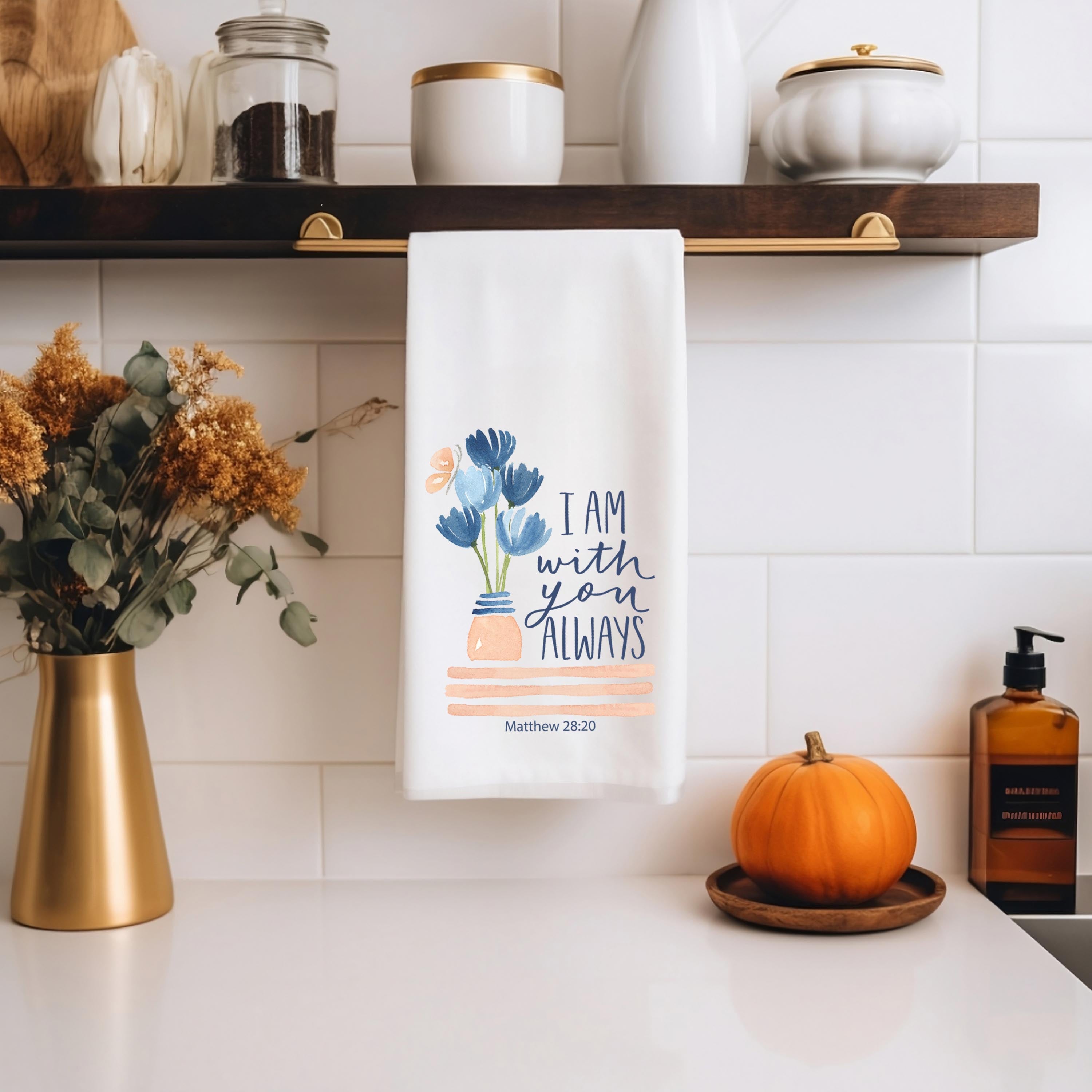 Decorative Christian kitchen towel with floral design and text "I Am With You Always" hanging on a kitchen shelf with pumpkins and decor items.