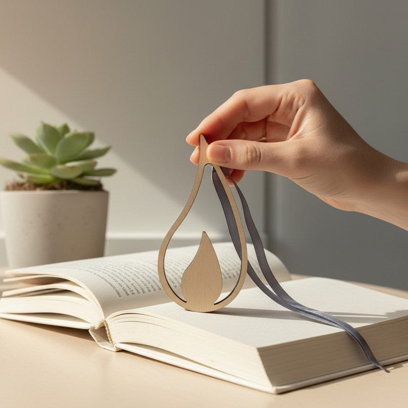 Hand holding a wooden flame object with a ribbon over an open book on a desk with plants in the background