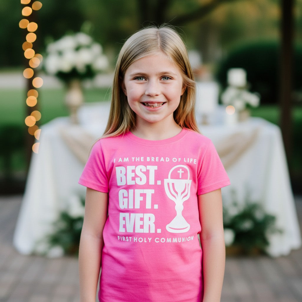 Young girl wearing a pink shirt with religious text for first communion, standing outdoors with blurred background