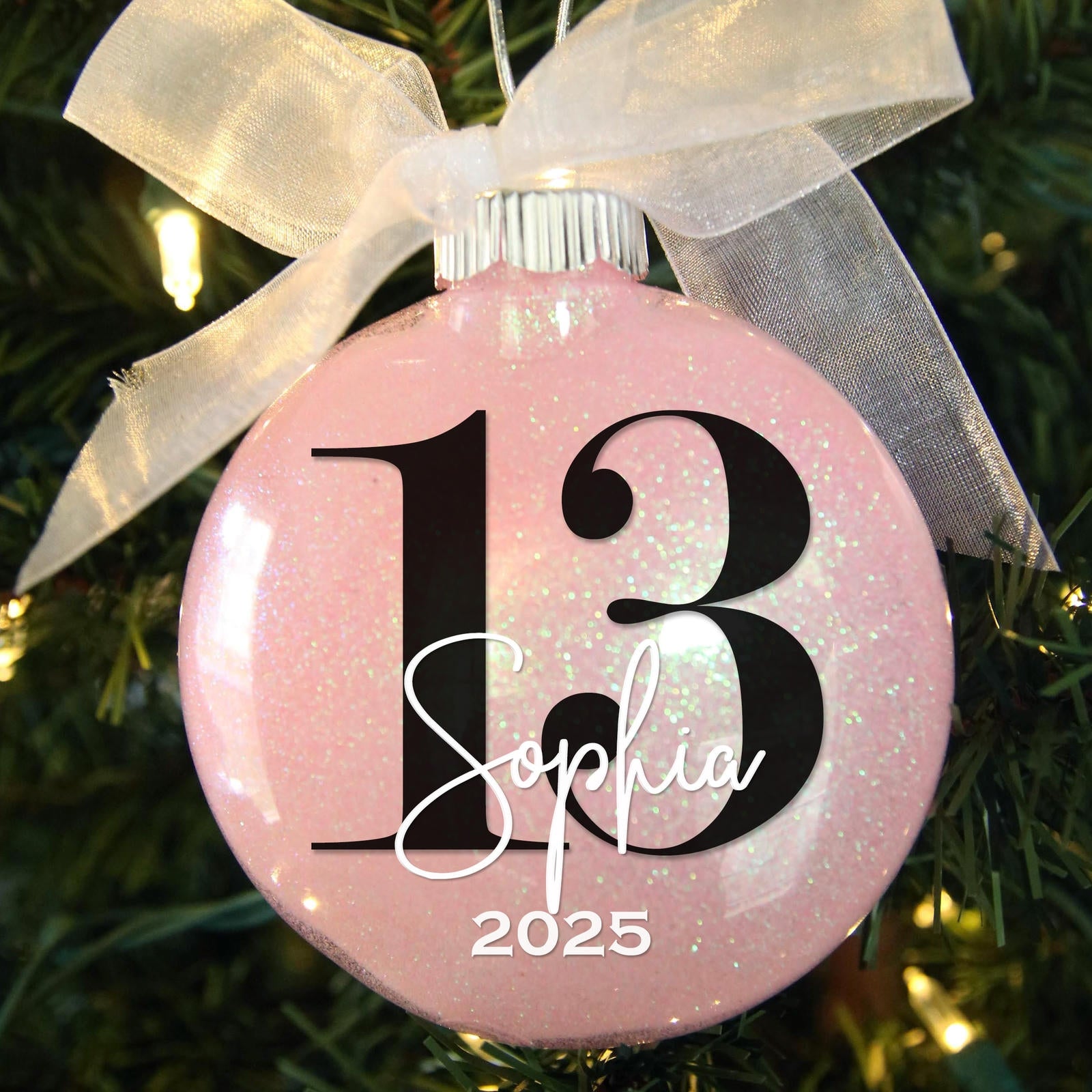 Light pink glittered ornament with the number 13 and Sophia 2024 hanging in a Christmas tree