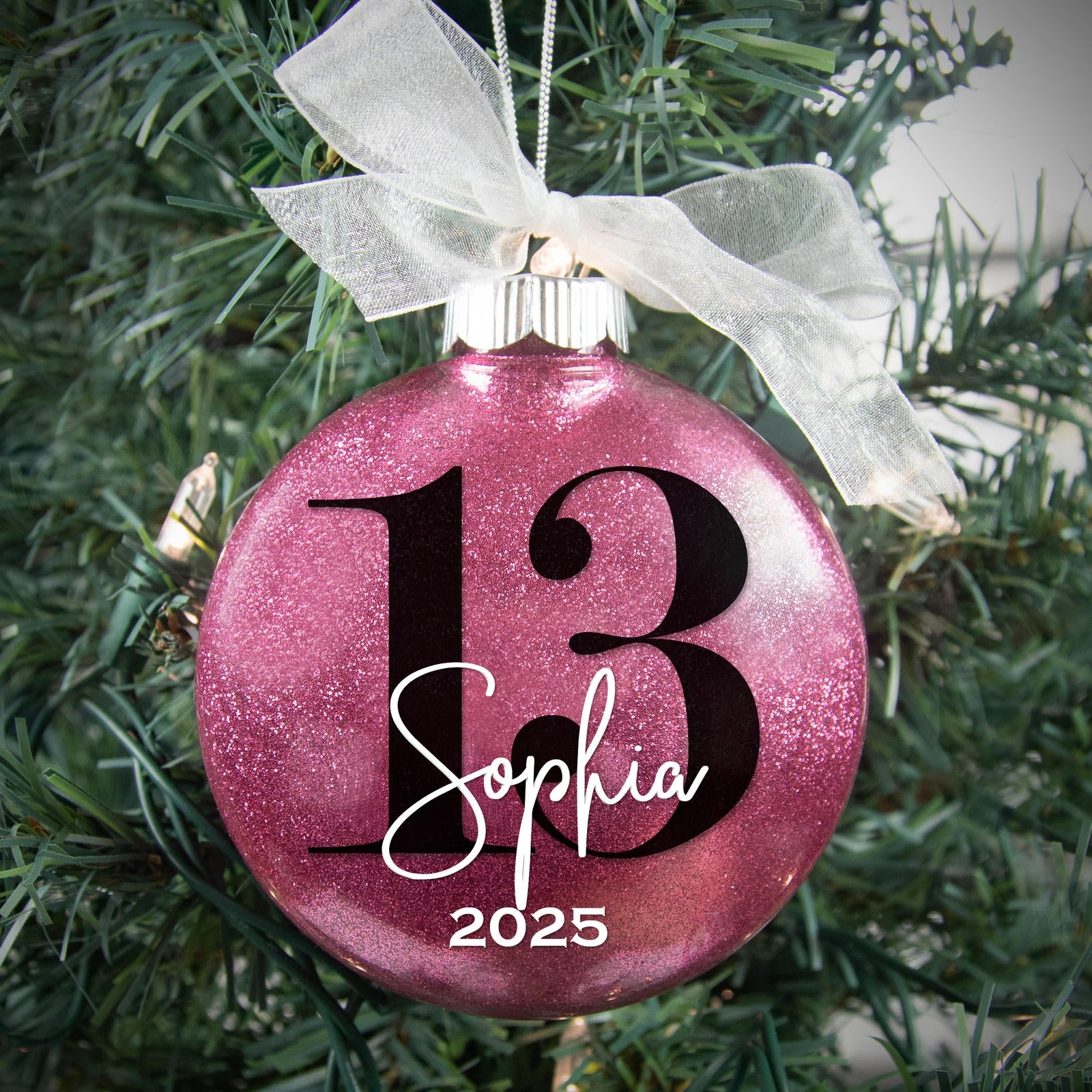 Raspberry colored glittered ornament with the number 13 and Sophia 2024 hanging in a Christmas tree